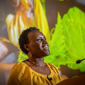 Dr Ngumbi speaking in a yellow shirt with plant presentation behind her